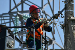 Electrical lineman in hard hat with face shield, hi-vis vest, and safety harness working on power transmission tower