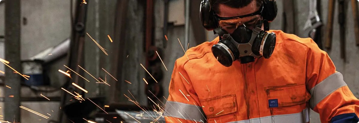 Worker in orange flame-resistant coveralls grinding metal with sparks flying, wearing respirator, goggles, and ear protection