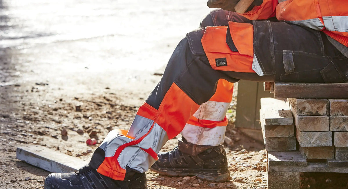 Worker wearing hi-vis work trousers with reflective tape and reinforced knees sitting on timber at construction site