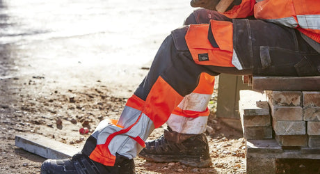 Worker wearing hi-vis work trousers with reflective tape and reinforced knees sitting on timber at construction site