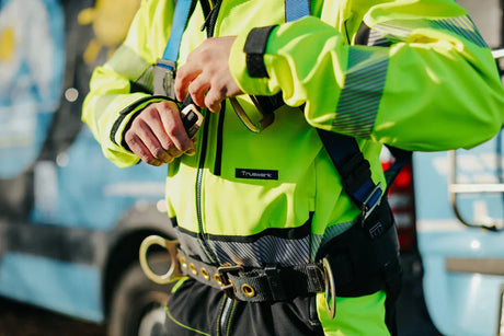 Worker adjusting fall protection harness over hi-vis shell jacket showing workwear layering system